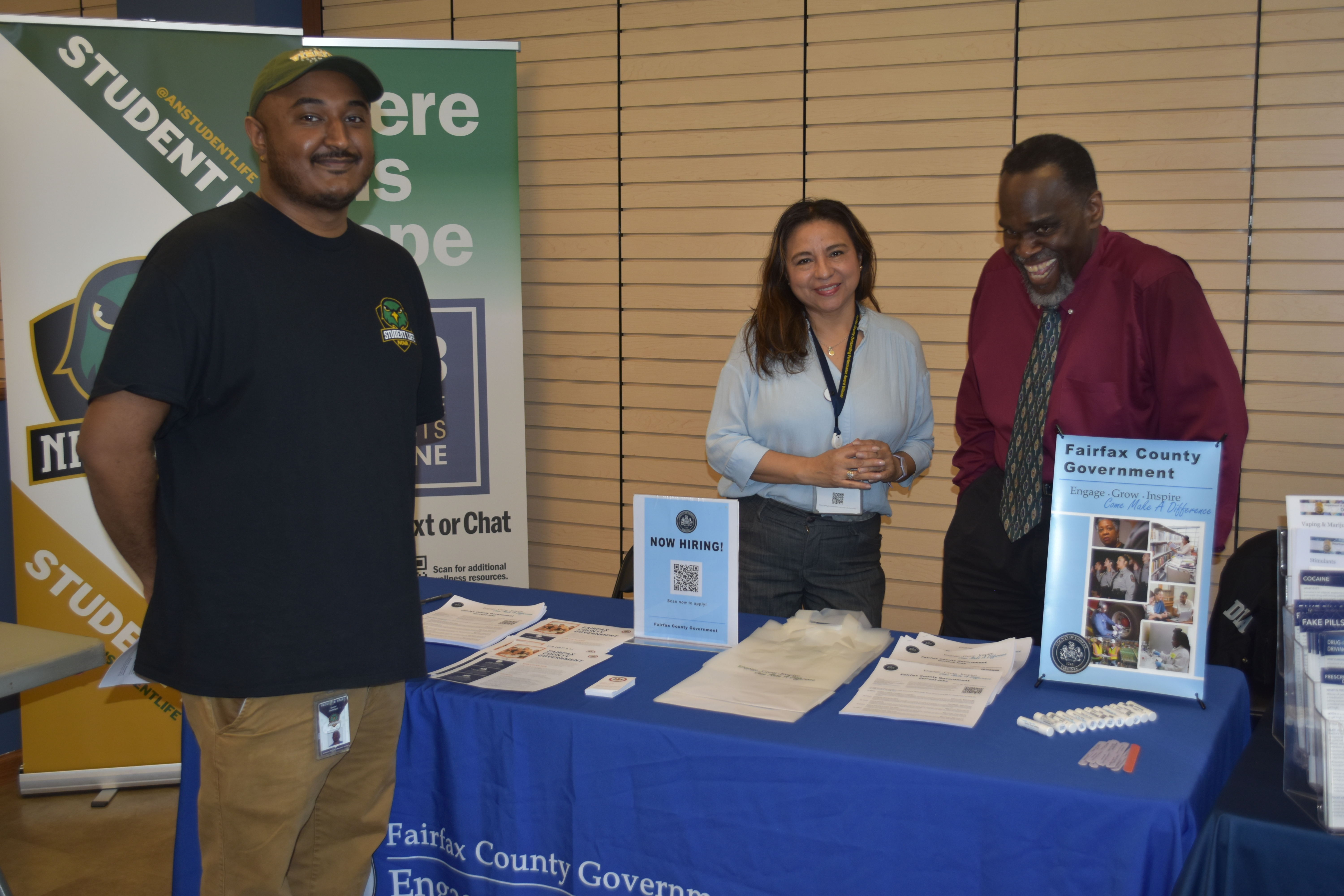 A Student Life representative and two Fairfax County Government recruiters standing behind a blue information table featuring 'Now Hiring' signs and career resources at a campus event.